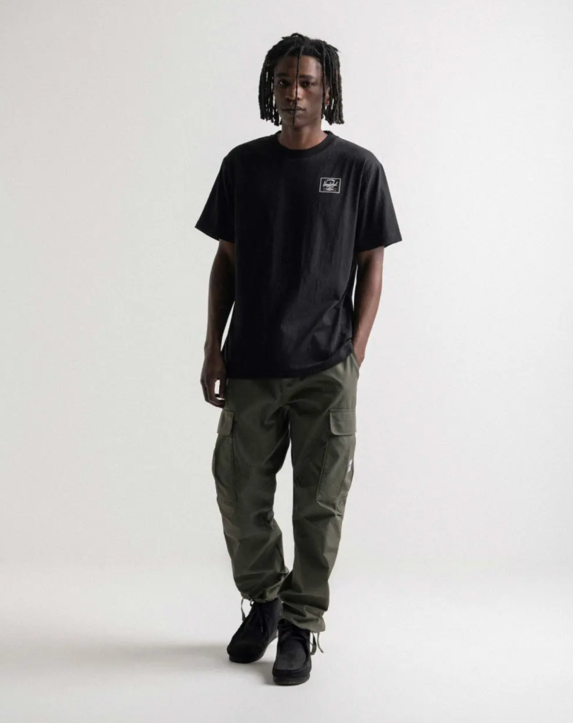 A man with dreads wearing a black Herschel logo t-shirt and green cargo pants