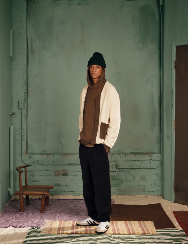 A man wearing a brown Kotn sweatshirt with a white cardigan over top of it along with a beanie, black pants and adidas shoes