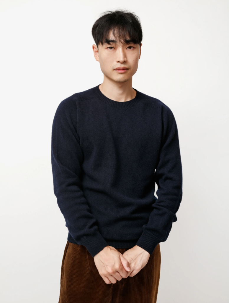 A man wearing a navy blue Neighbour sweatshirt and brown pants on a white backdrop