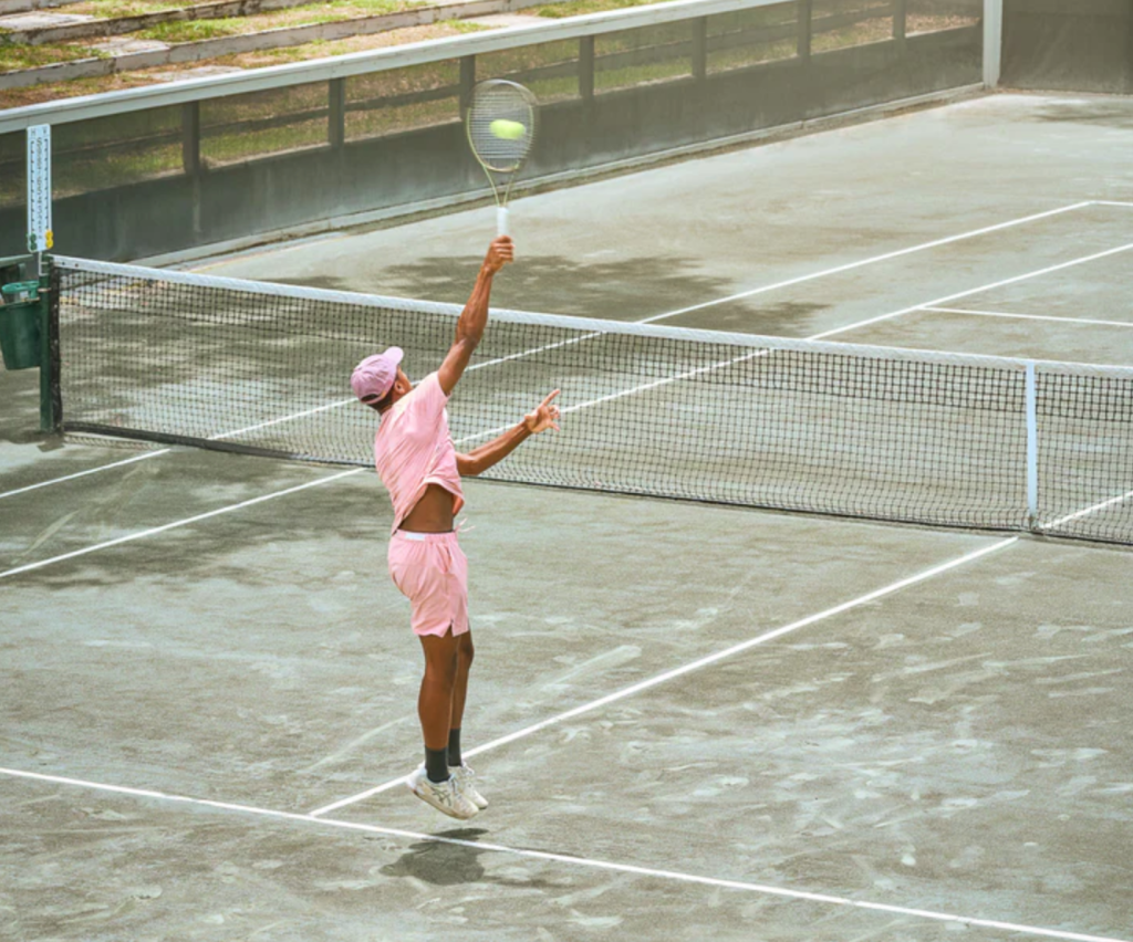 A man serving on a tennis court while wearing a pink Sigrun tennis outfit
