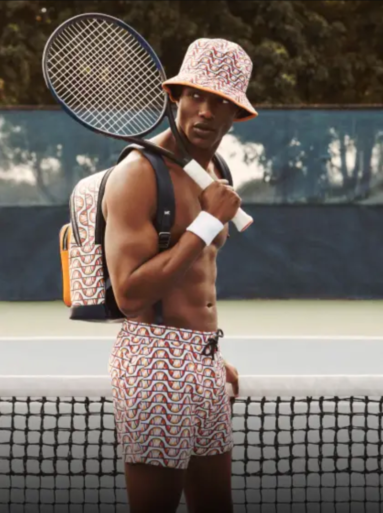 A man with a tennis racket over his shoulder wearing matching colorful Elesse shorts, backpack and bucket and no shirt