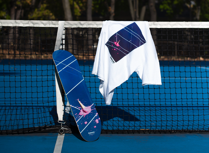 A white t-shirt with a tennis court print on it draped over a tennis court net with a matching skateboard balanced beside it