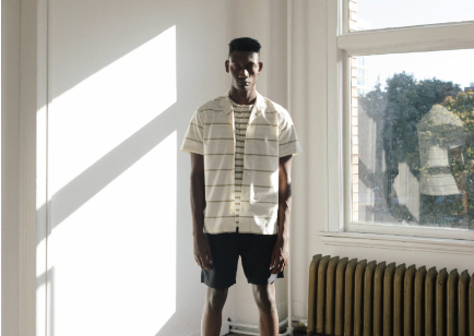 A man in a sunlit room wearing a Wings+Horns short sleeve striped button up and navy blue shorts