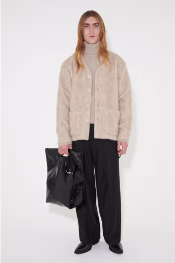 A man wearing a beige Stephan Schneider turtleneck and a beige cardigan over top along with black pants and bag