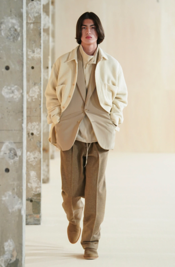 A man with long hair wearing a beige Auralee overshirt on top of a light brown blazer along with a turtle neck & brown pants