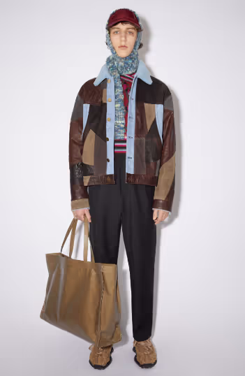 A man wearing an all over printed Acne Studios jacket with a colorful sweater underneath along with a scarf and brown bag