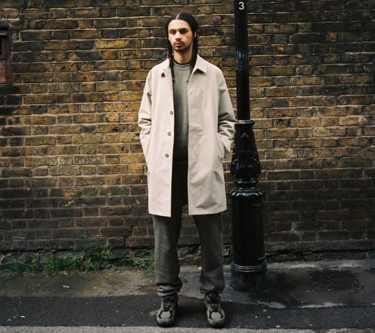 A man standing on the street wearing a biege JJJJound trench coat, green sweatshirt, dark grey pants and shoes