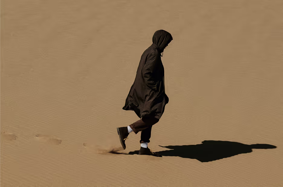 A man walking in the sand with a black Nanamica jacket wearing his hood with black pants, white socks and black boots on