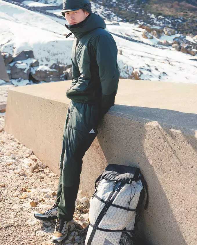 A man wearing a green And Wander snow suit with a matching green hat and salomon shoes