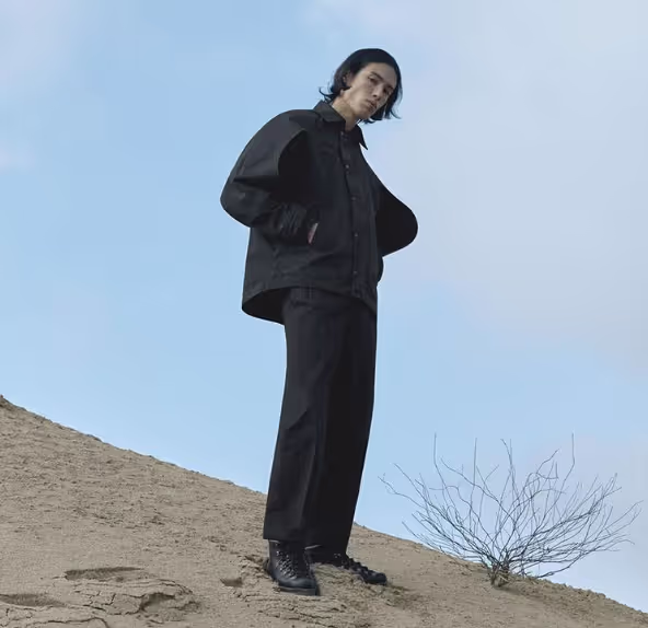 A man standing on a sandy hill wearing a black Konundrum jacket with matching black pants and boots