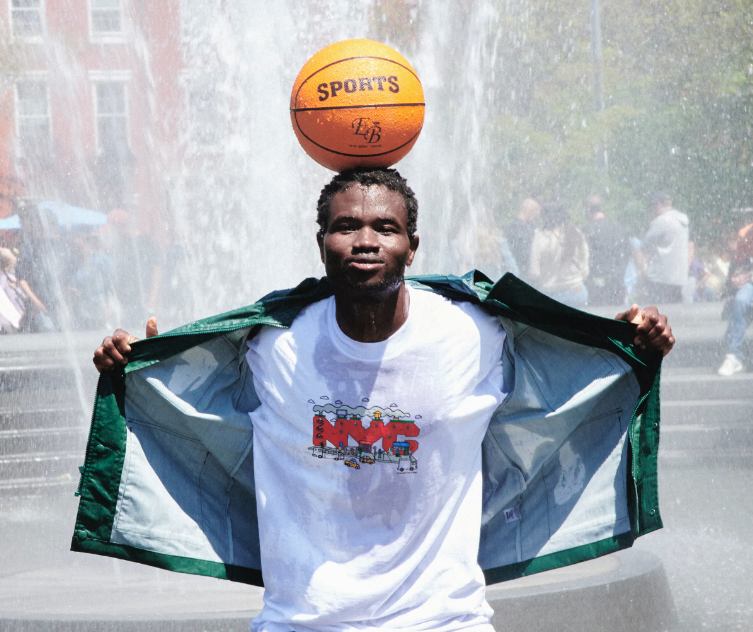 A man standing in a sprinkler with a basketball balanced on his head wearing Only NY t-shirt and jacket