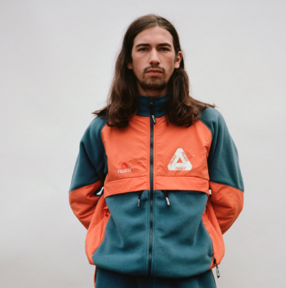 A man with long brown hair wearing an orange and green 90s style Palace fleece jacket