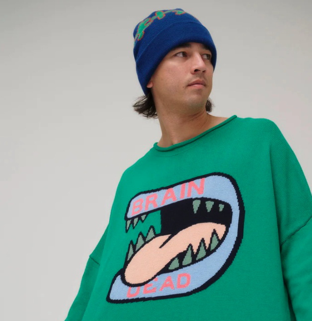 A man wearing a green Brain Dead Sweater with a drawing of a monsters mouth with it's tongue out on it and a blue beanie