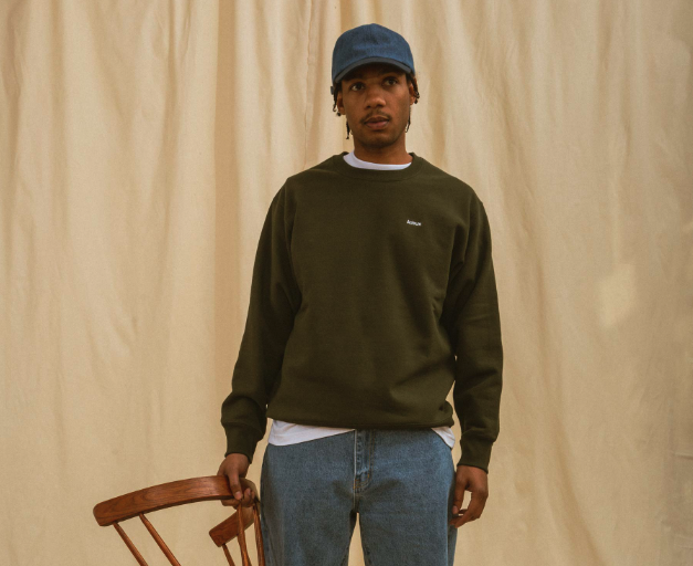 A man wearing a green Adsum sweater, jeans and a blue denim hat