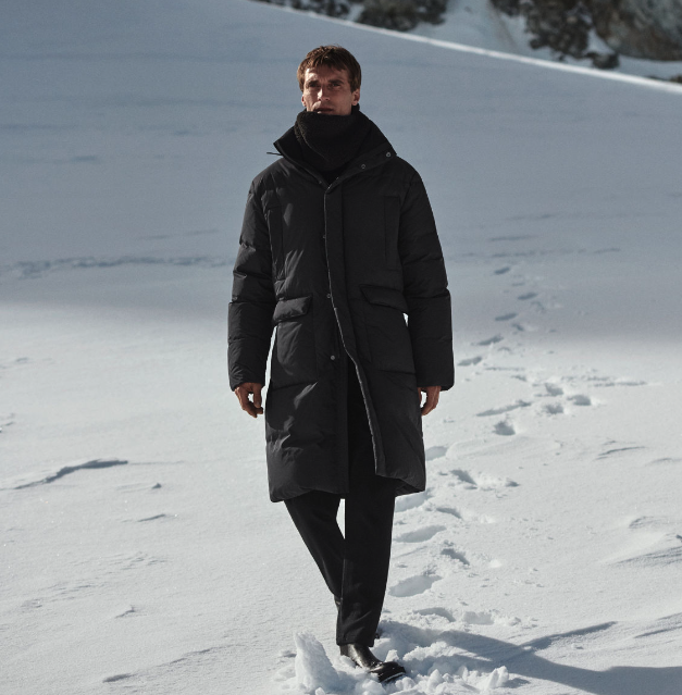 A man bundled up wearing a dark grey COS jacket, scarf and pants walking in the snow