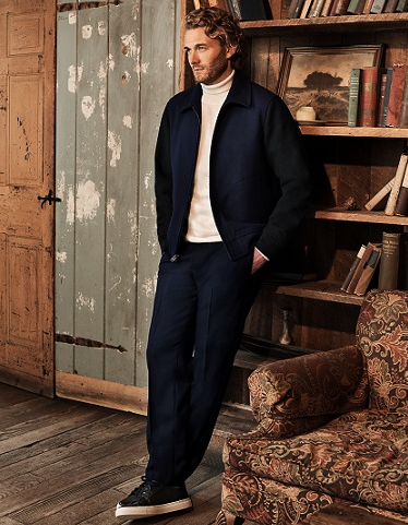 A man with blonde curly hair in a lodge cabin waring a navy blue Club Monaco jacket and pants, with a beige turtleneck