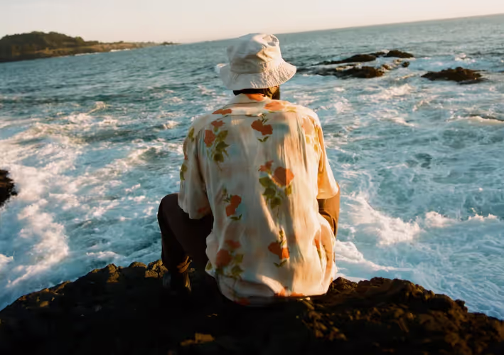 A man sitting while looking out into the ocean wearing a floral General Admission button up and a white bucket hat