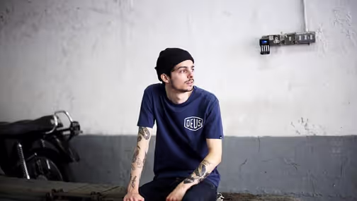 A man looking off into the distance wearing a Navy Blue shirt with a large Deus logo and matching beanie