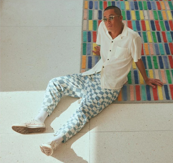A man sitting on the floor wearing light blue and white Banks Journal checkered pants, a beige button up and sunglasses
