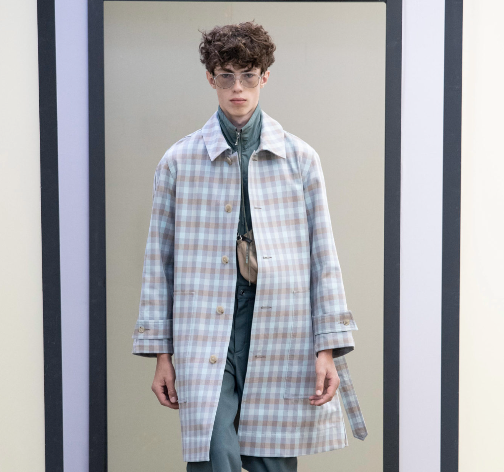A man wearing a baby blue checkered print Maison Kitsune trench coat and clear vintage glasses