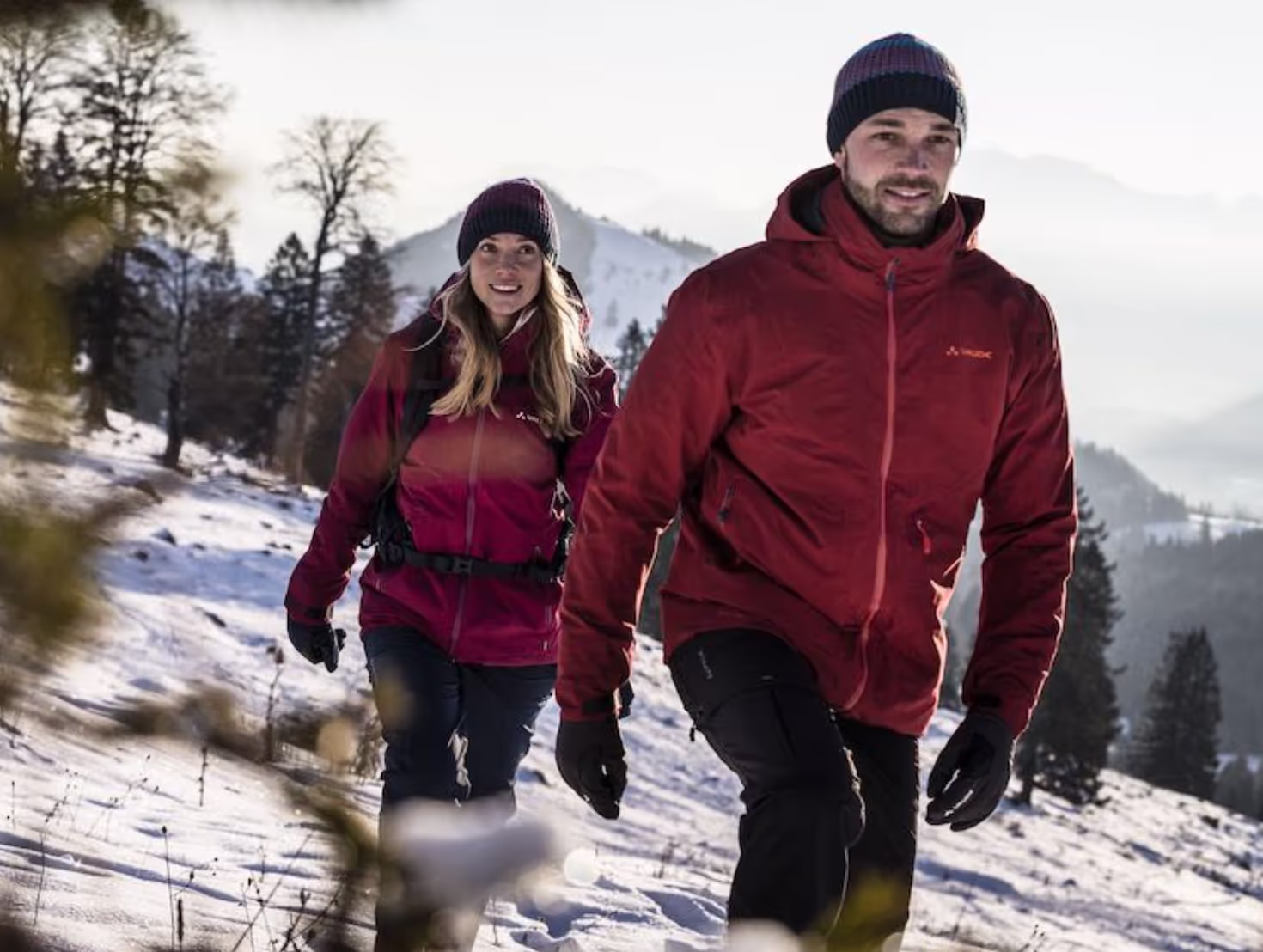 A couple hiking in the snow wearing matching Maroon Red Vaude Jackets, black pants and a matching beanies