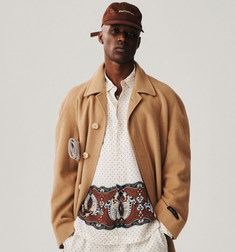 A man wearing a brown Aime Leon Dore trench coat and white button up with a dark brown hat