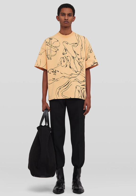 A man wearing a peach colored Jil Sander illustrated short-sleeved t-shirt with black pants and black boots