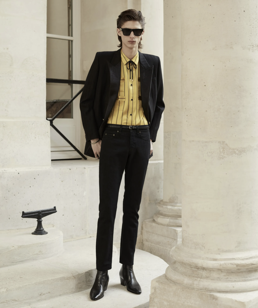A slim man wearing a Yves St Laurent yellow striped button up with a bolo tie, black blazer, pants and shoes.