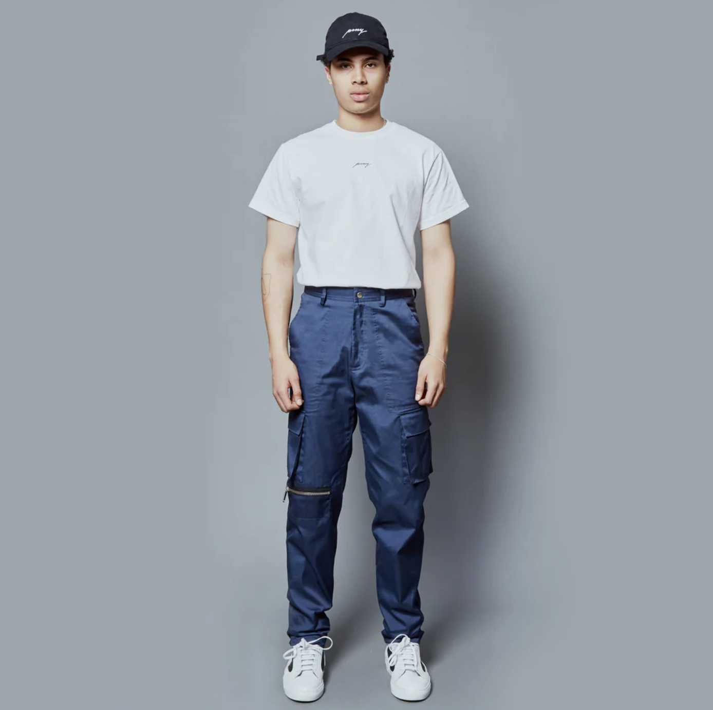 A man wearing a Public School NYC white logo t-shirt and navy blue hat and pants along with white sneakers