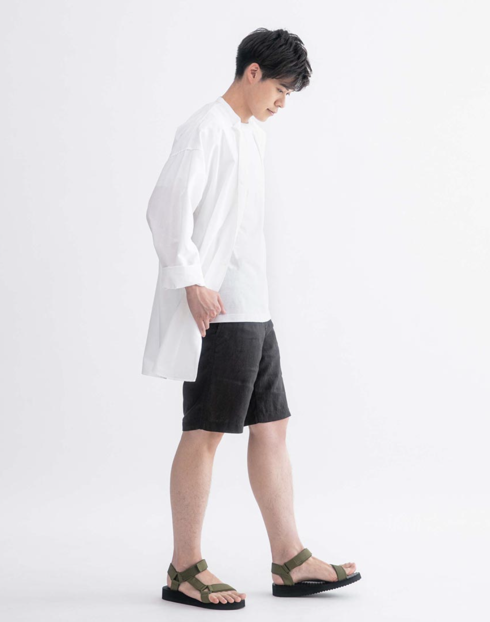 A man wearing a long white Muji button up along with black shorts and olive sandals