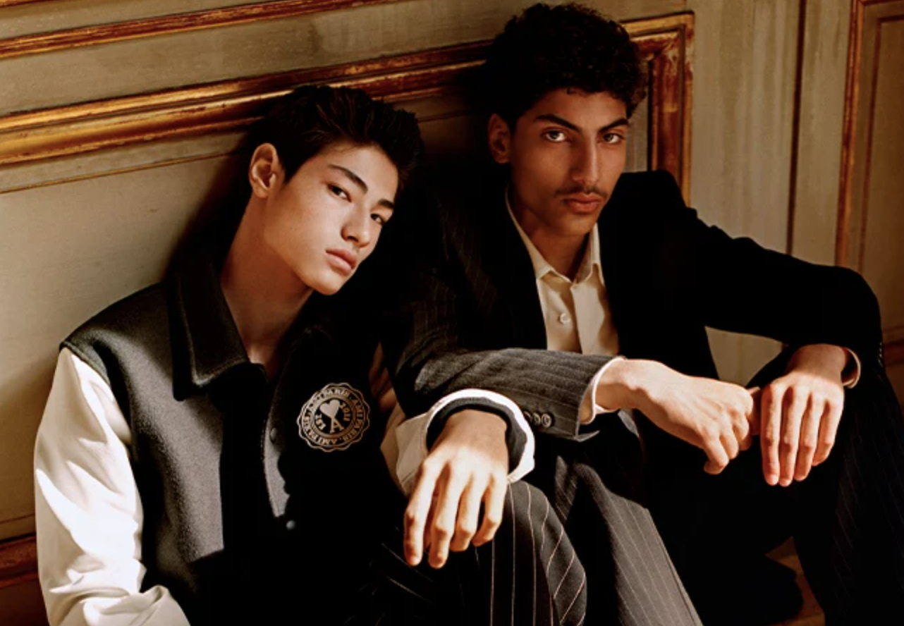 Two men sitting together wear Ami outfits featuring a varsity jacket and suit