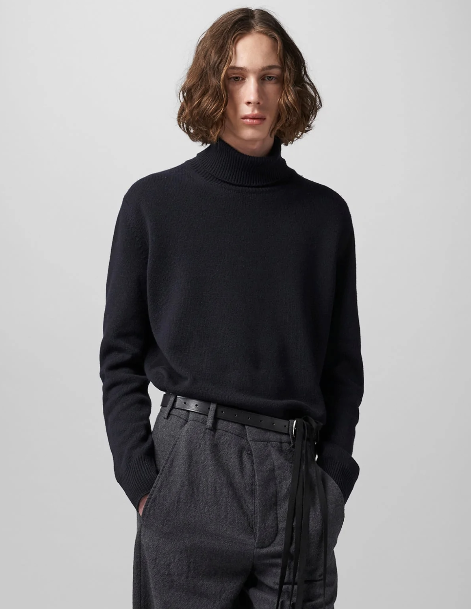 A man wearing a black Ann Demeulemeester turtleneck with a belt and dark grey dress pants