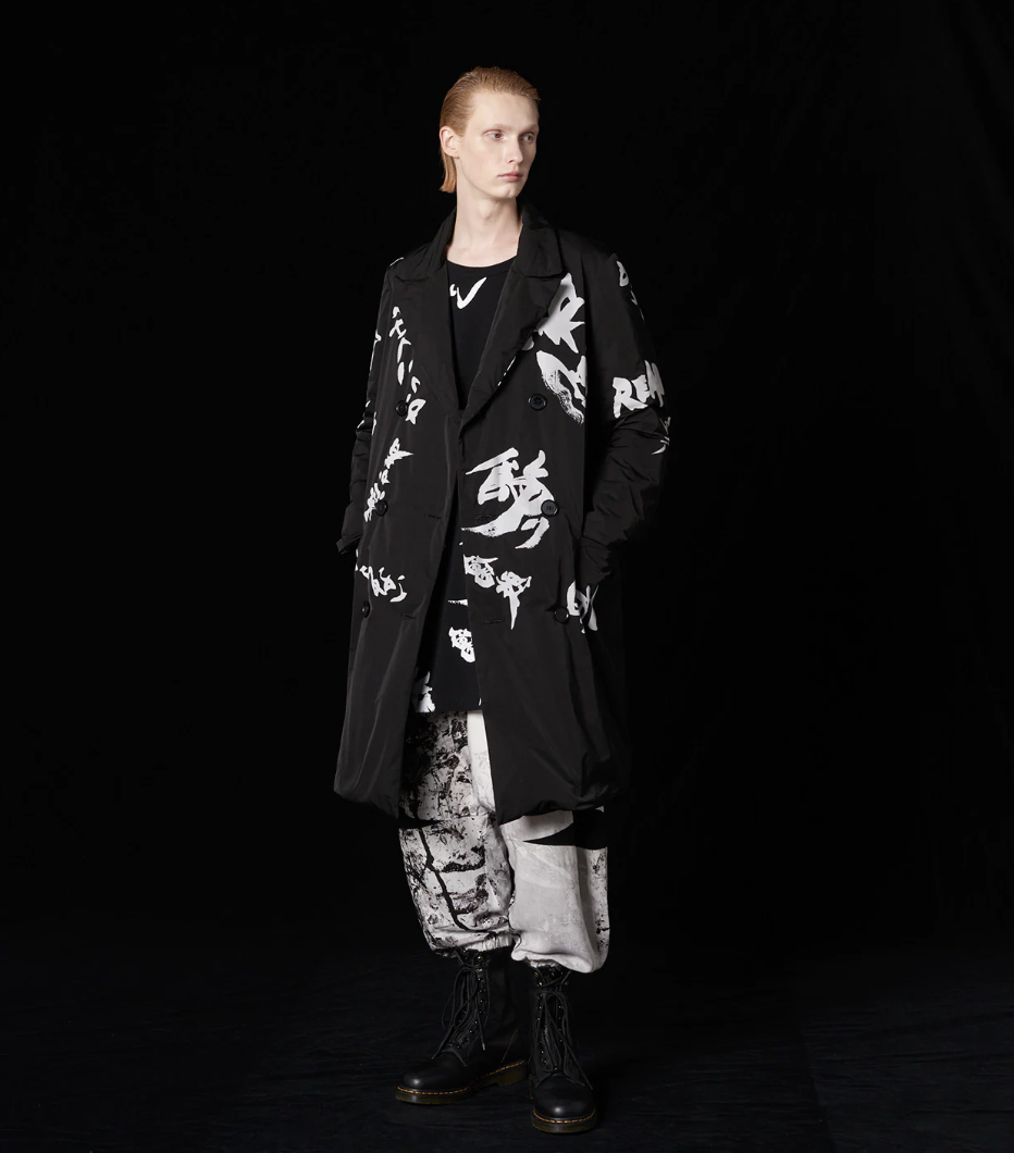 A man with long slicked back hair wearing a Yohji Yamamoto black trenchcoat with white brush strokes across it and black boot