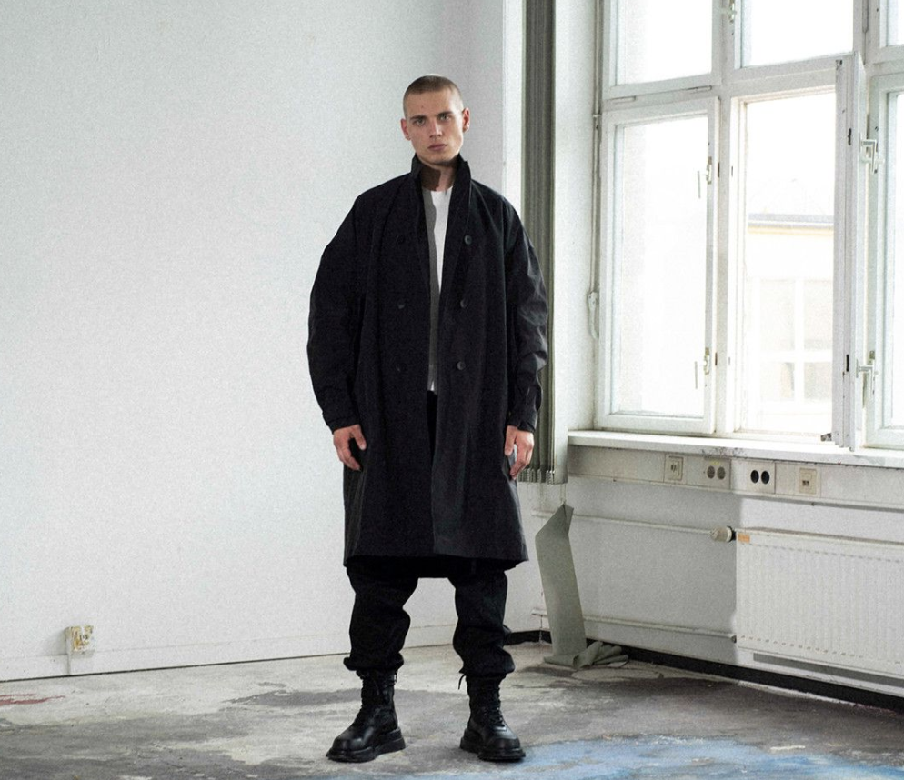 A man wearing a Julius outfit feating a black trenchcoat, black pants and boots along with a white shirt