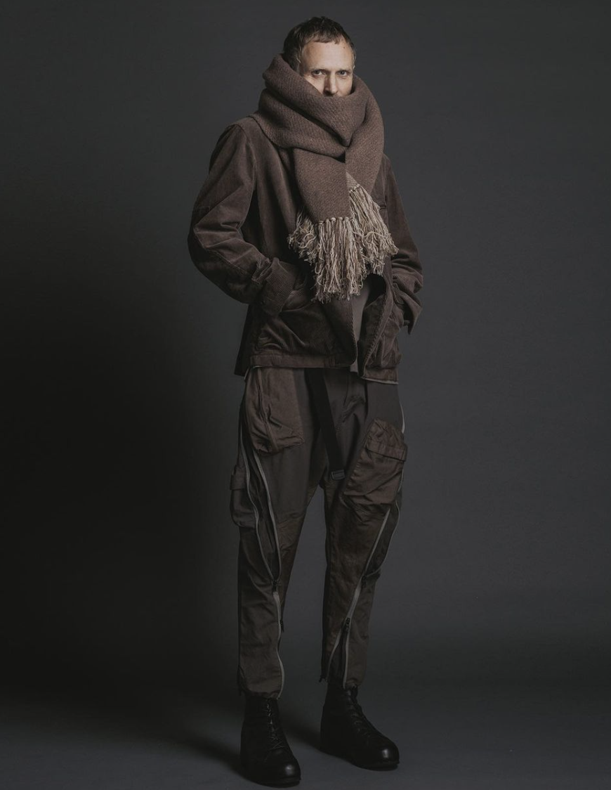 A man wearing The Viridi-Anne oversize brown scarf, a brown jacket with technical looking pants and black hi-top sneakers