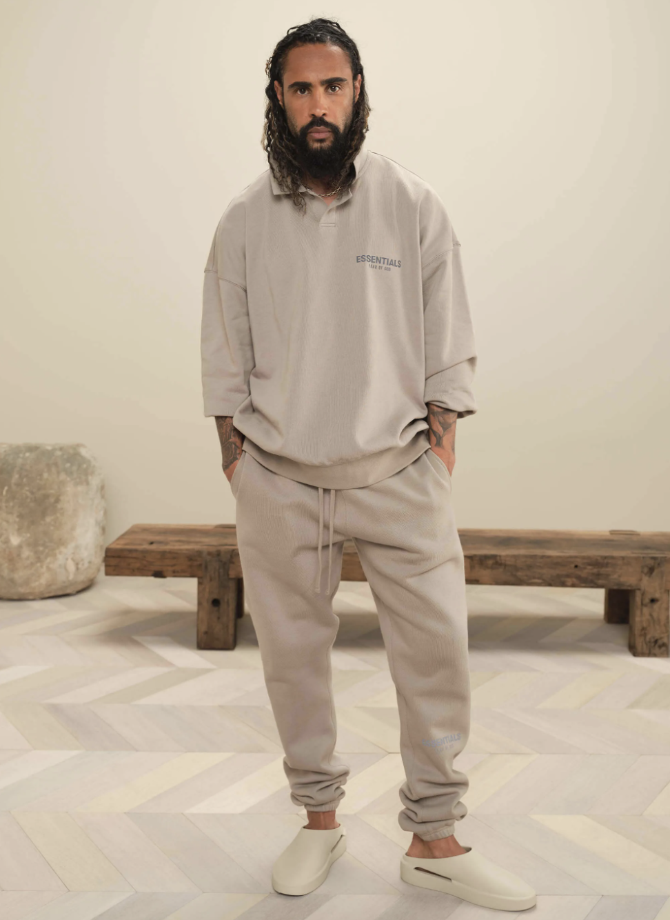 A man wearing Fear of God Essentials loungewear including a beige long-sleeve, matching sweatpants and cream slippers