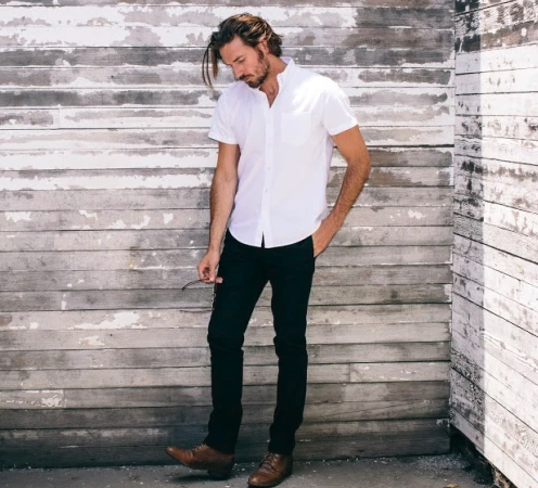 A man wearing a Buck Mason white button-up short-sleeve with black pants and brown loafers