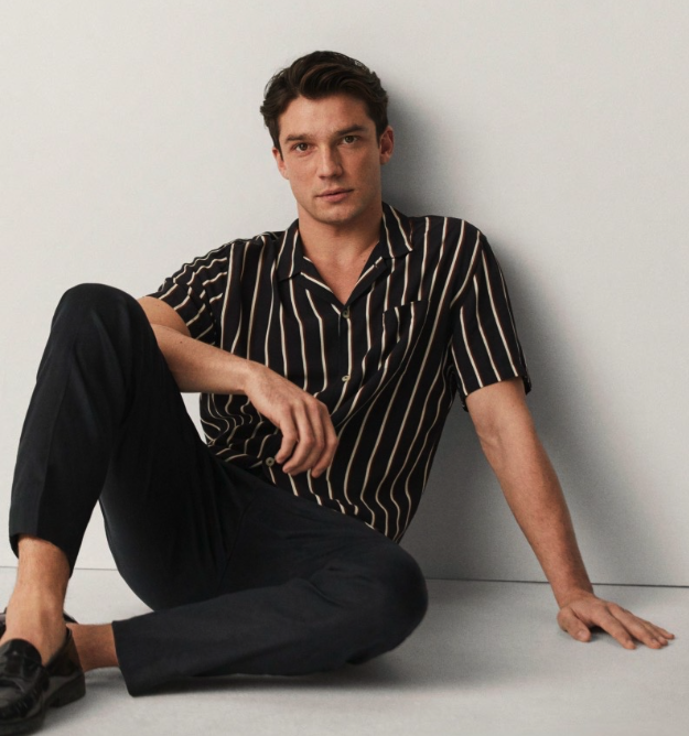 A man sitting against a wall wearing a Mango striped short-sleeve black button up and black tapered pants