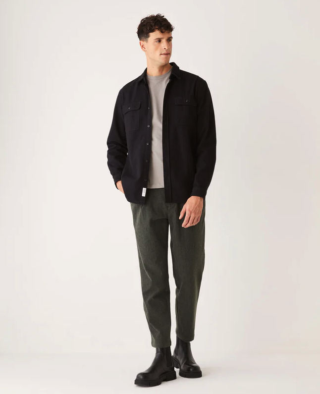 A man wearing a black Frank & Oak button up shirt with dark green pants and black boots