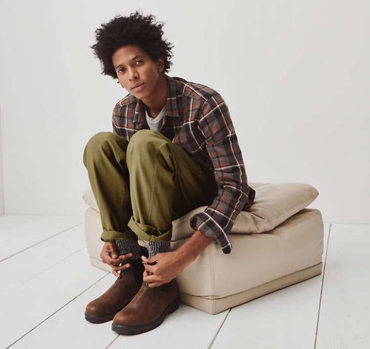A man sitting and wearing a Madewell plaid button up, green pants and brown blundstone boots