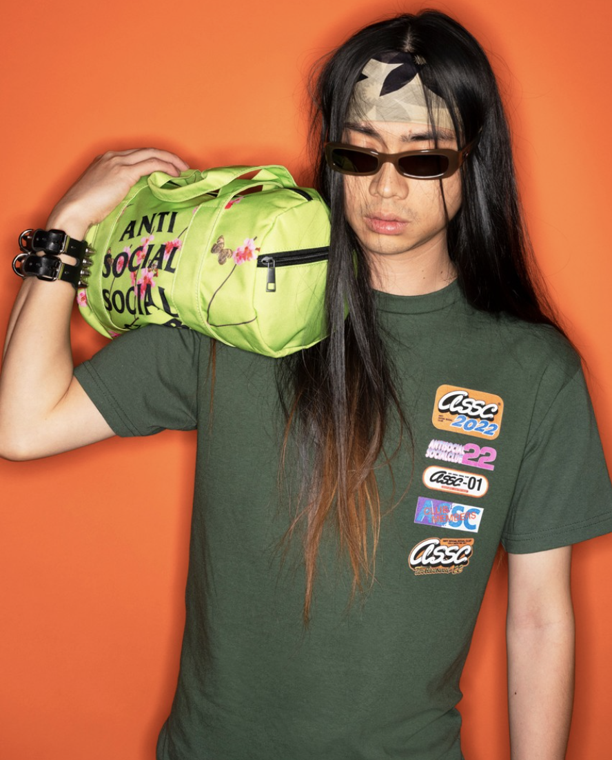 A man with long hair wearing a green Anti Social Social Club t-shirt while holding a lime green small duffle bag