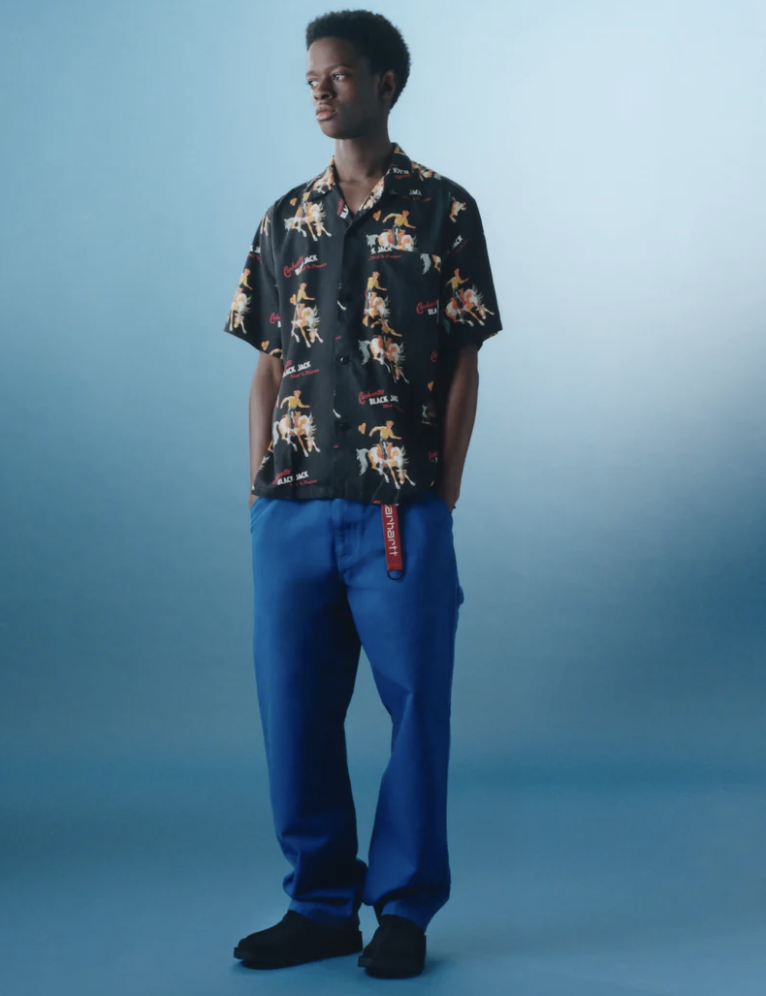 A man wearing a Carhartt WIP black graphic button-up with blue pants and branded carhartt belt