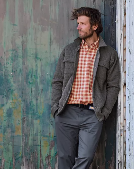 A man leaning against a wall wearing an Orvis plaid shirt and overcoat with workwear pants