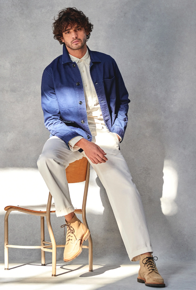 A man sitting on a chair while wearing a Todd Snyder outfit with a denim overcoat and cream pants