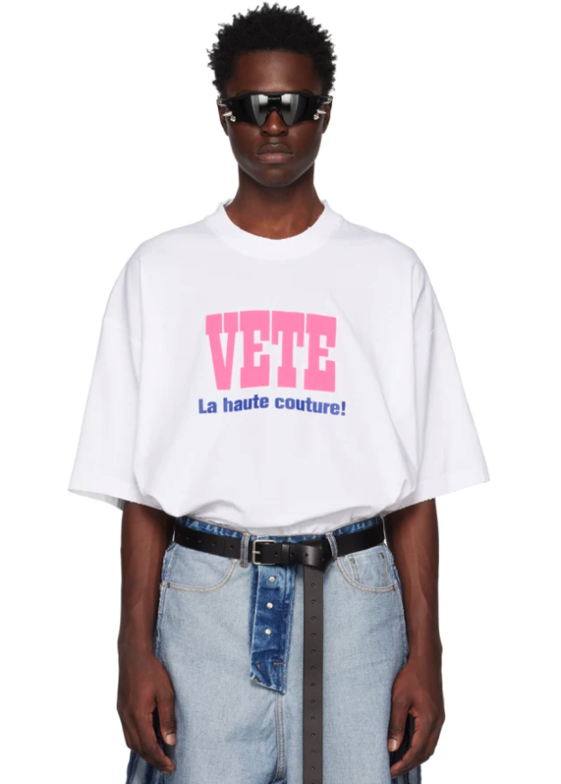 A man wearing a Vetements outfit featuring a white VETE t-shirt and blue jeans