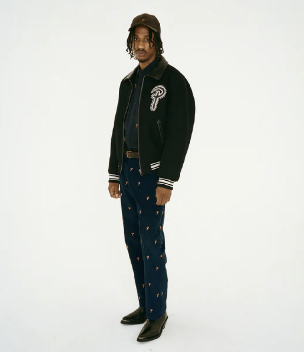 A man wearing a Patta coaches jacket with navy blue pants and black pointed dress shoes