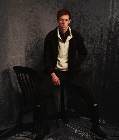 A man wearing a dark Mr. Oliver jacket, beige zip sweater with black pants and boots