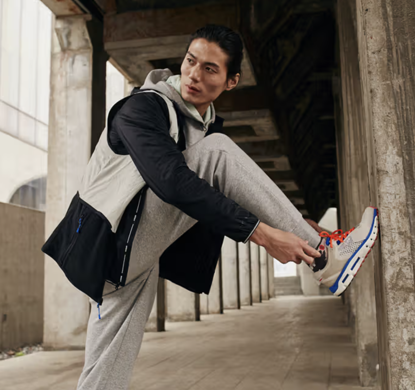 A man tying up his Under Armor shoes while he wears an Under Armor grey sweatpants and a black and grey jacket
