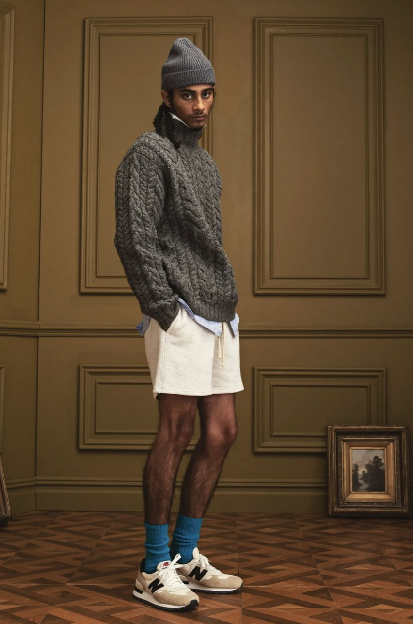 A man in a New Balance outfit with a dark grey fisherman sweater, a matching beanie and blue socks