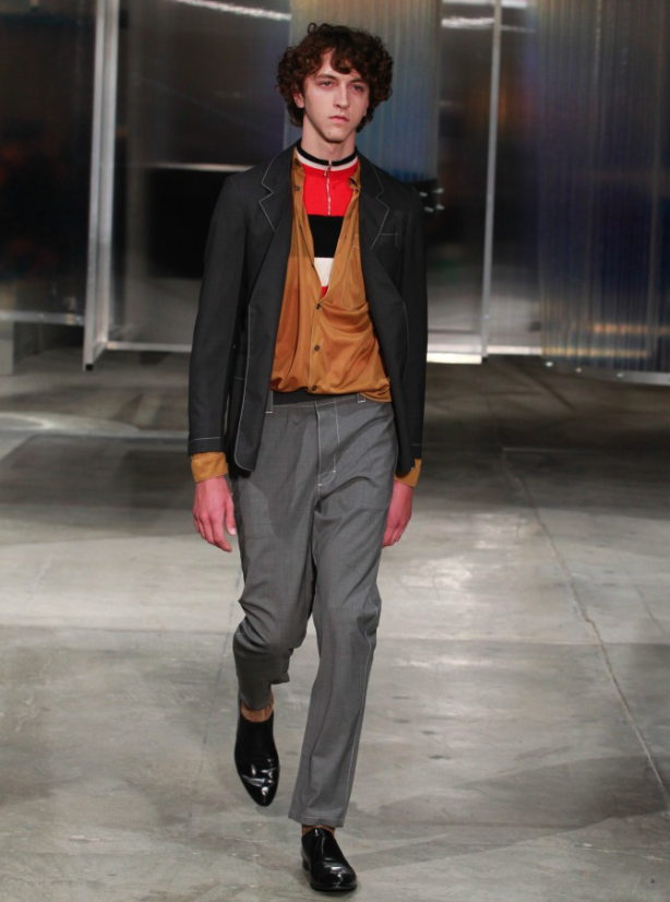 A man wearing a Prada outfit with grey pants and a brown cardigan